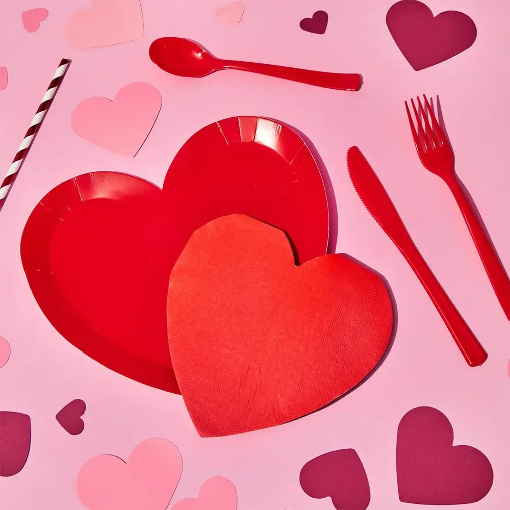 Red heart-shaped paper plates with a matching napkin, surrounded by red plastic cutlery, a striped straw, and scattered pink and red paper hearts on a pink background.